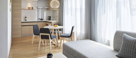 A modern hotel room with a grey sofa, a dining table with chairs, a kitchen area, and a large window with curtains.