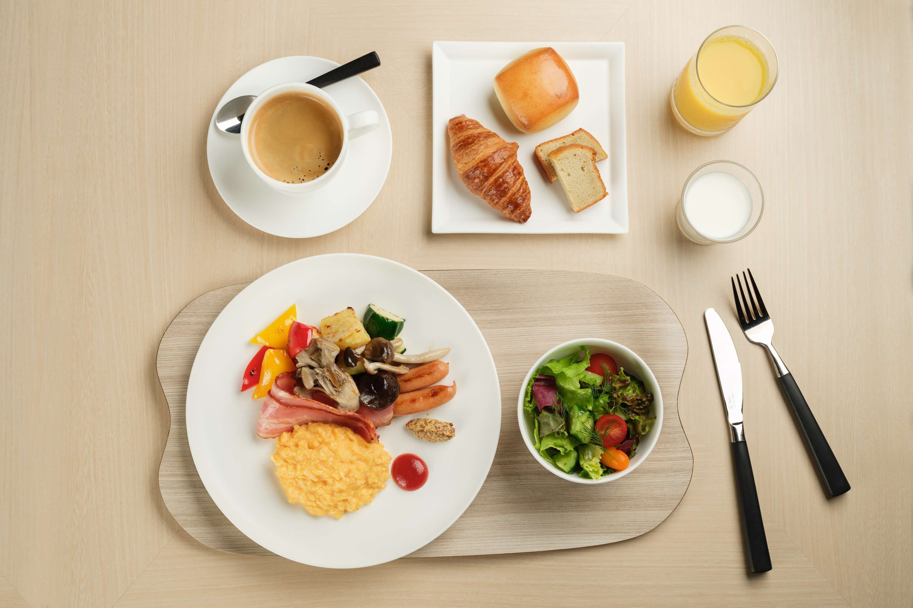 Une table dressée pour le petit-déjeuner avec une assiette d’œufs brouillés, du jambon, des légumes, un croissant, des toasts, un saladier, une tasse de café, un verre de jus d’orange et des couverts, le tout posé sur une table en bois.