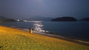 A beach at night with a visible moon, calm water, and distant lights.