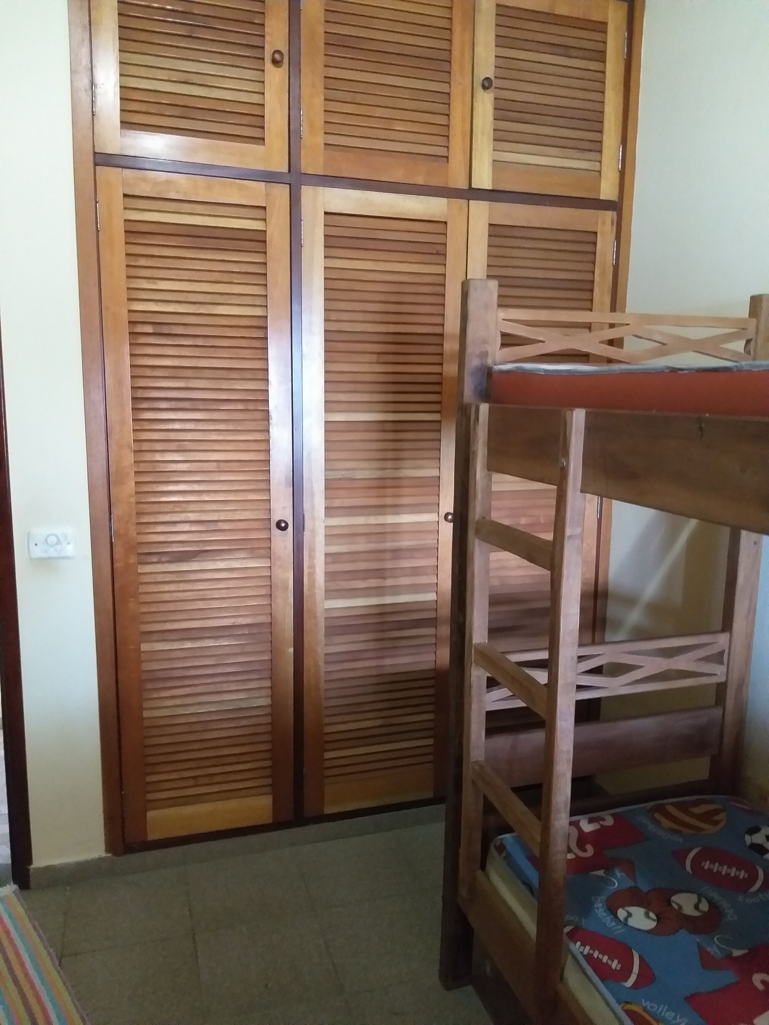 Uma cama beliche com colchão estampado, um guarda-roupa de madeira com ripas horizontais e um chão ladrilhado.