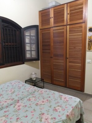 A bedroom with a bed, a wooden wardrobe, a small table with a cup, and a window with shutters. - House in Massaguaçu walk-in-the-sand! (caraguatatuba)