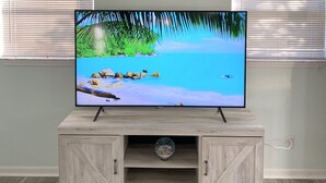Ein Flachbildfernseher auf einem Holz-TV-Möbel mit einer Urlaubsansicht eines tropischen Strandes, vor einer hellen Wand mit zwei Fensterläden.