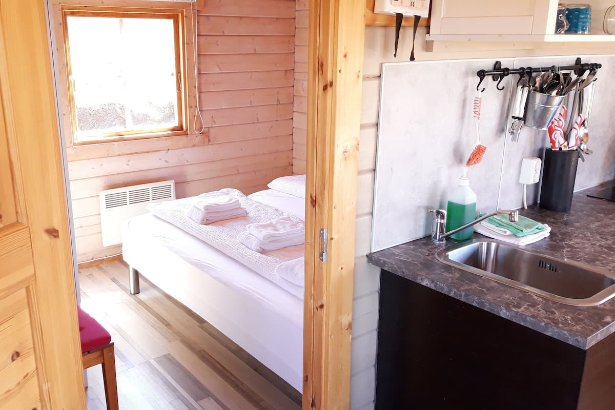A wooden cabin interior with a bed, a small kitchenette, and a window.