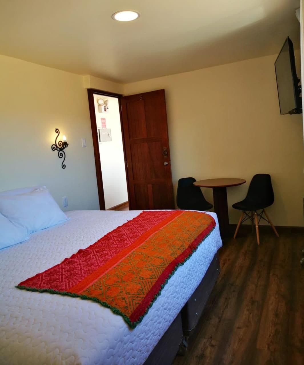 A hotel room with a bed, a small round table, two chairs, a wooden door, and a wall-mounted light fixture.