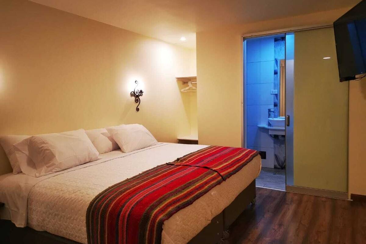 A bedroom with a bed, a bedside lamp, a wardrobe, and a bathroom visible through an open door.