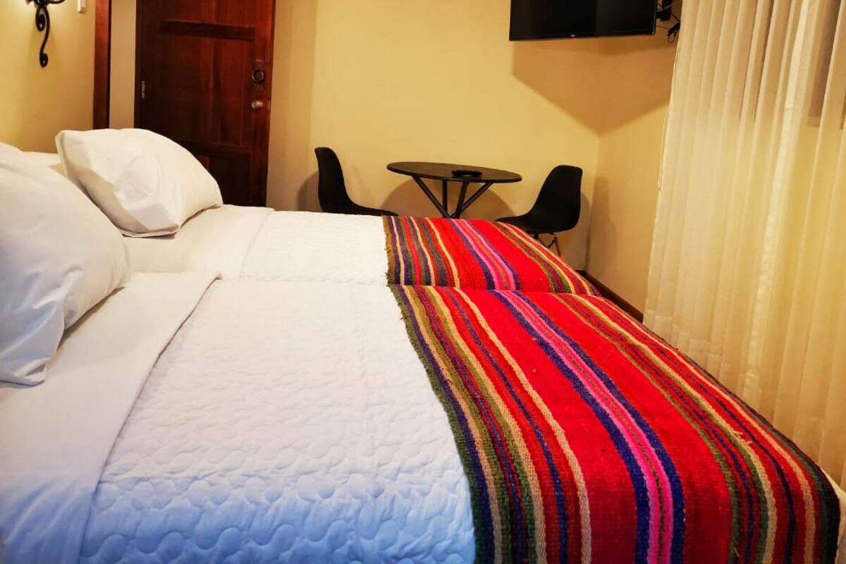A hotel room with a bed, a striped blanket, a small table, two chairs, a TV, and a window with curtains.