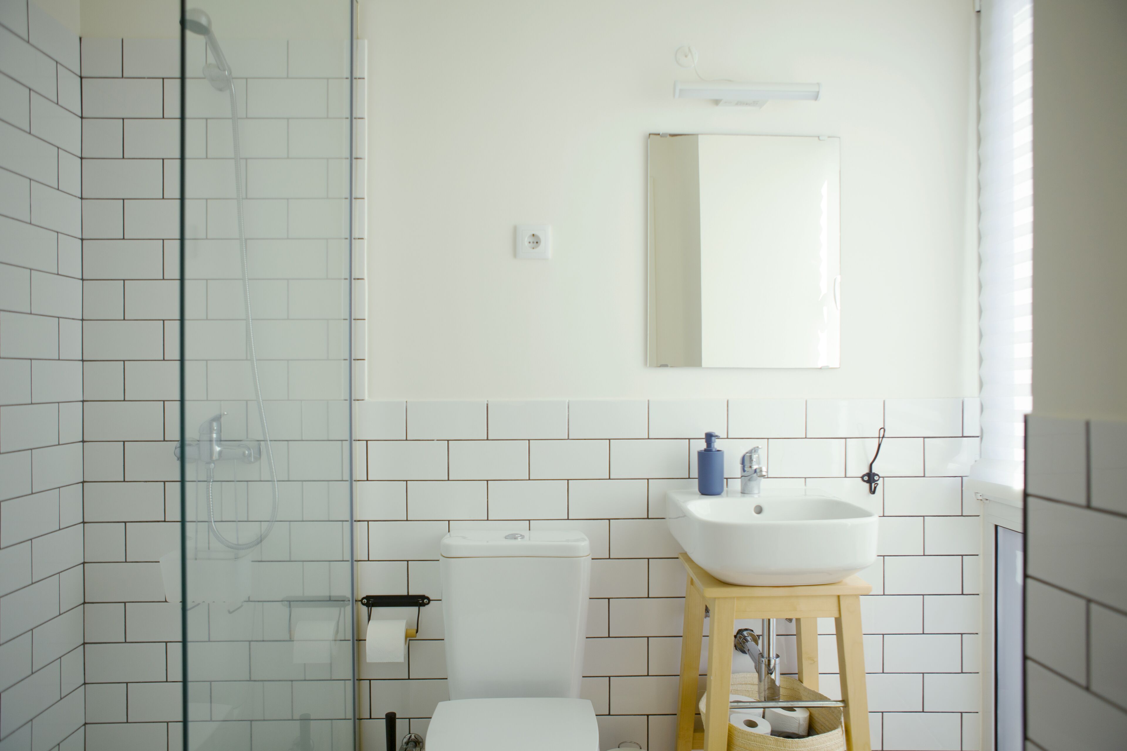 Une salle de bain avec une douche à parois de verre, des toilettes blanches, un lavabo avec un tabouret en bois et un miroir au mur.
