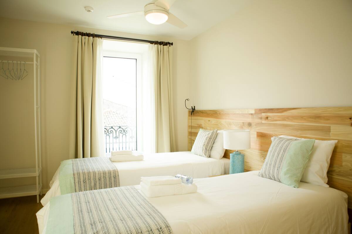 A hotel room with two beds, a ceiling fan, a window with curtains, and a wooden headboard.
