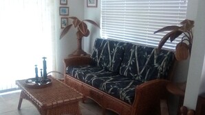 Olohuoneessa on sohva, sohvapöytä ja lattiavalaisin. Seinällä on tauluja ja ikkunassa sälekaihtimet.