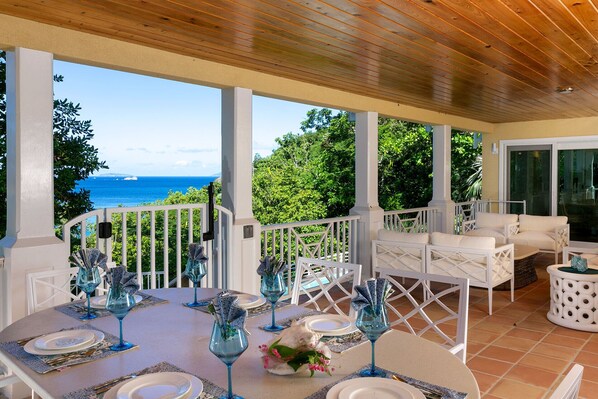 A covered outdoor dining area with white furniture, a table set for four, and a view of the ocean. - Kallaloo: Walk to Gibney Beach! (St. John)