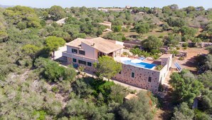 A two-story stone house with a swimming pool, surrounded by dense greenery and hills.