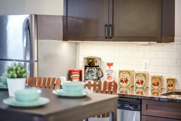 A modern kitchen with stainless steel appliances, a wooden dining table with light blue plates, and various coffee and sugar boxes on the counter.