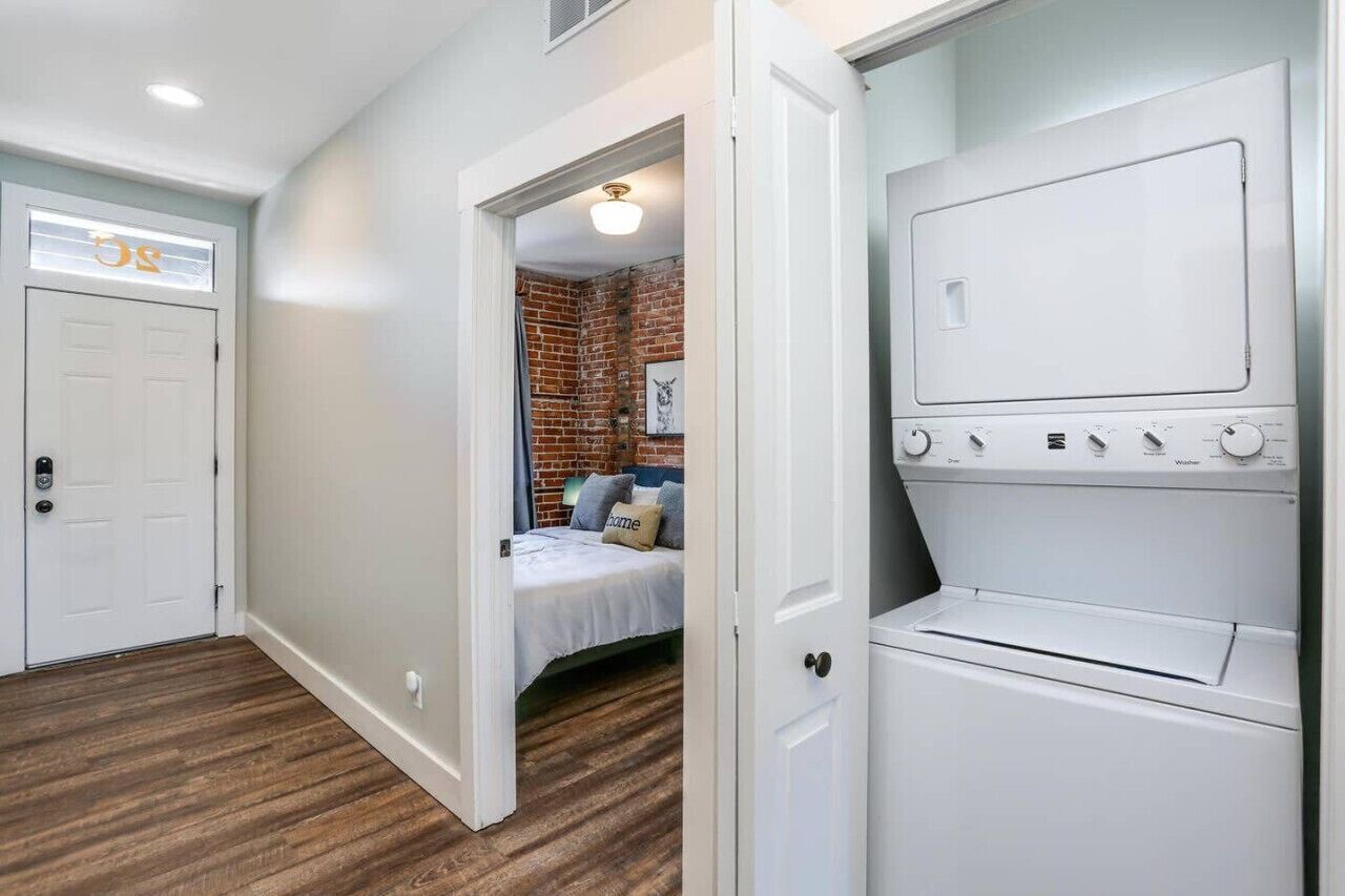 Uma lavanderia com máquina de lavar e secadora brancas, piso de madeira e porta que dá para um quarto com parede de tijolos e cama.