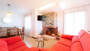 A living room with a brick fireplace, a television mounted above it, and a dining area with wooden furniture. - Sa Marina by dracmallorca (Cala Santanyi)