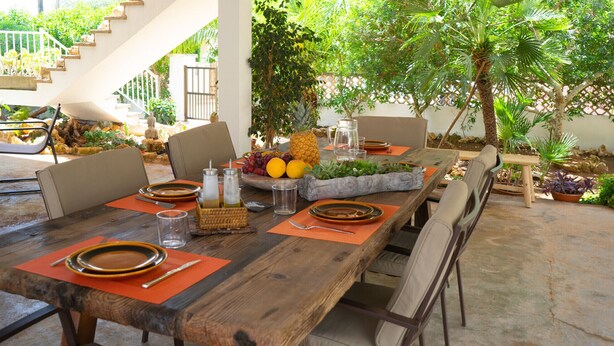 An outdoor dining area with a wooden table set for four, surrounded by chairs. There are potted plants and a small fountain in the background. - Sa Marina by dracmallorca (Cala Santanyi)