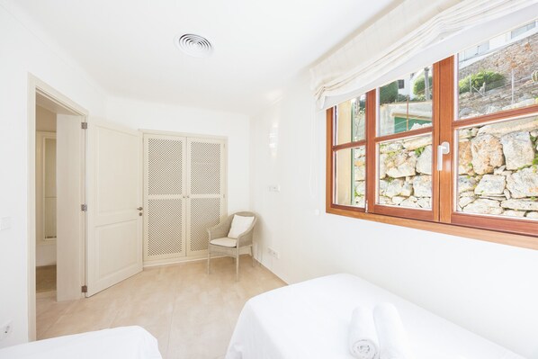 A room with a bed, a chair, a wardrobe, and a window with a stone wall view.