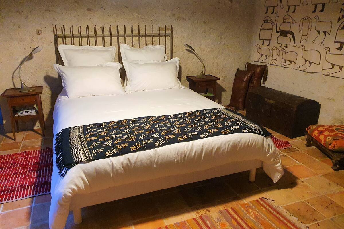A bed with white linens and a patterned blanket, a wooden headboard, two bedside tables with lamps, a small rug, and a wall with a mural.