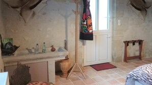 A room with a rustic stone wall, a wooden door, a small window, a coat rack with a colorful garment, a wooden chair, and a tiled floor. - Les Voies Blanches (Vouvray)
