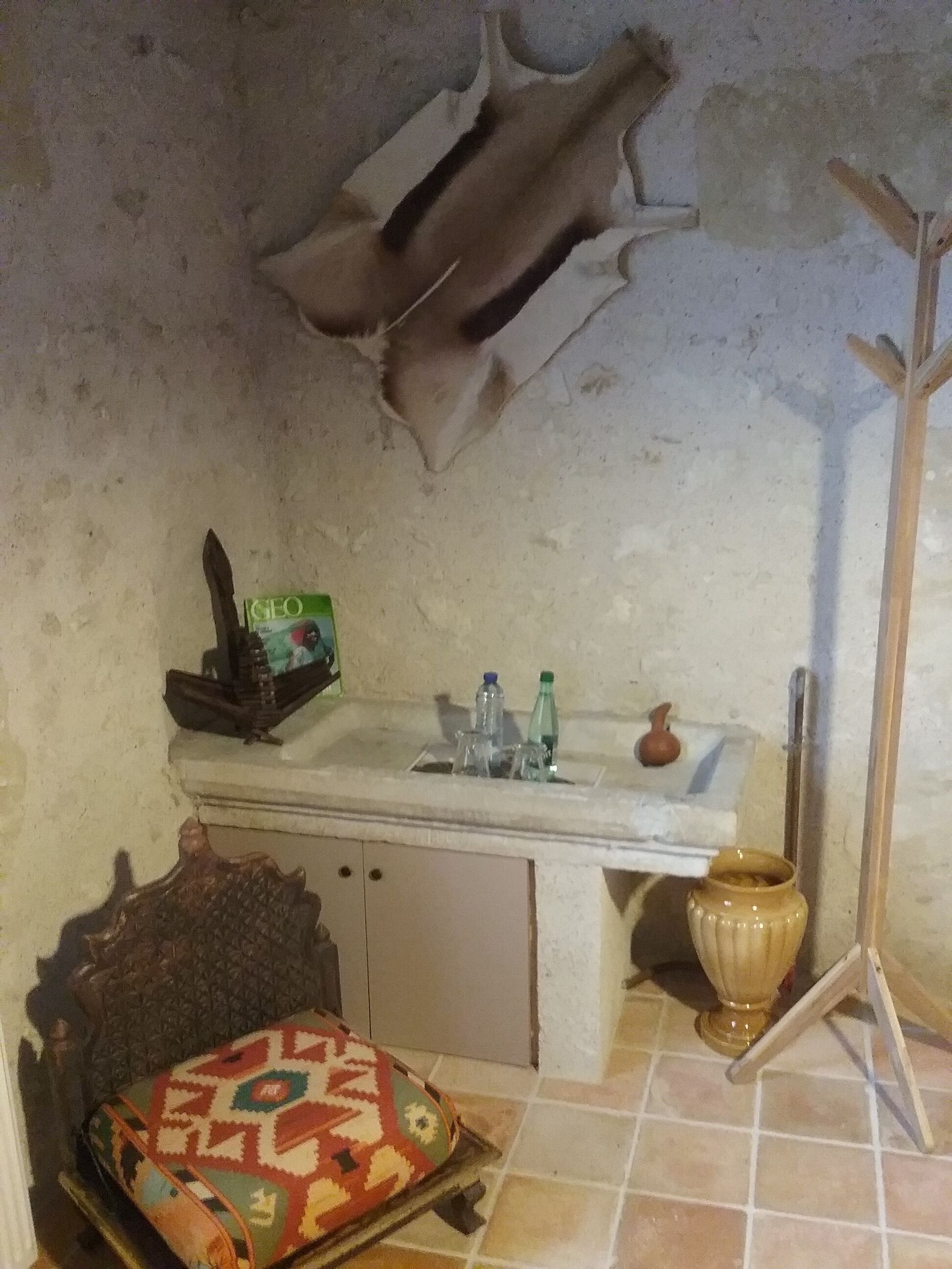 Una habitación pequeña y rústica con una cama individual, una silla de madera, una mesita con una botella y un estante de pared con una caja etiquetada “GEO”.