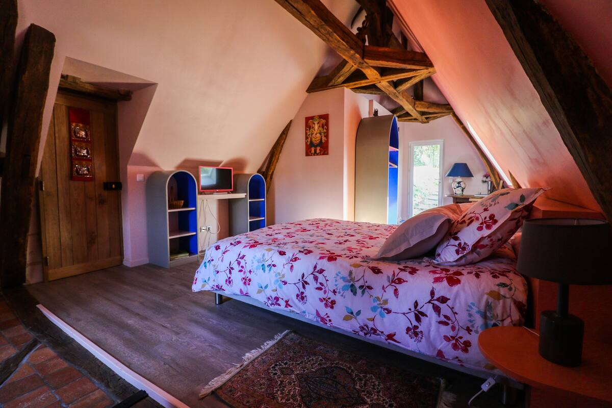 A bedroom with a sloped ceiling, a bed with floral bedding, a wooden door, and a small table with a lamp.