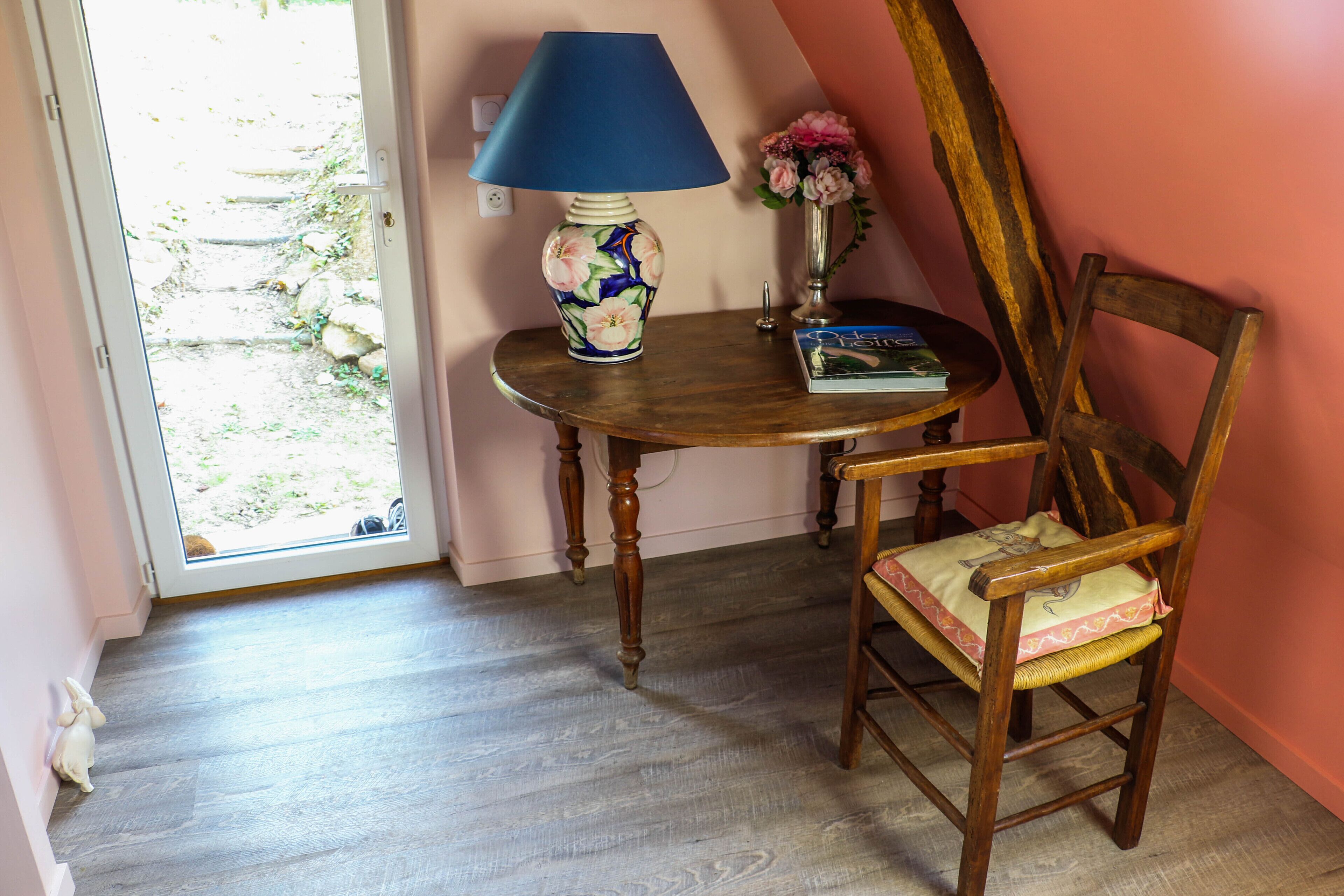 Quarto com cadeira de madeira, mesa com abajur azul, vaso com flores e um livro.
