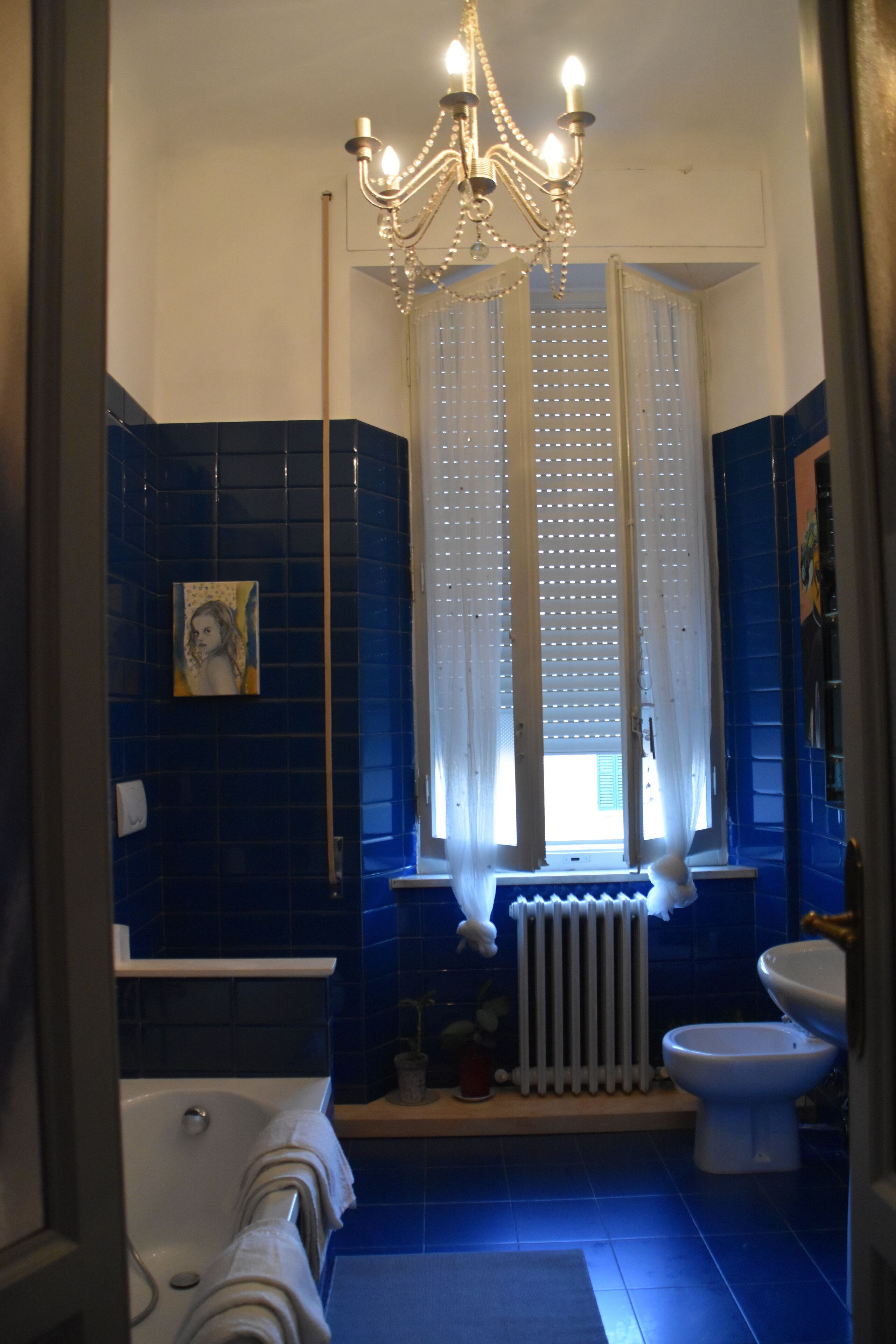 A bathroom with a bathtub, a toilet, a sink, a chandelier, and a window with curtains.