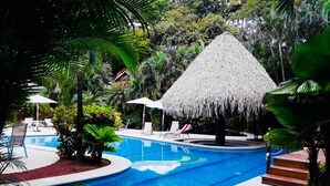 Una piscina con sillas de descanso y una estructura con techo de paja rodeada de plantas tropicales.