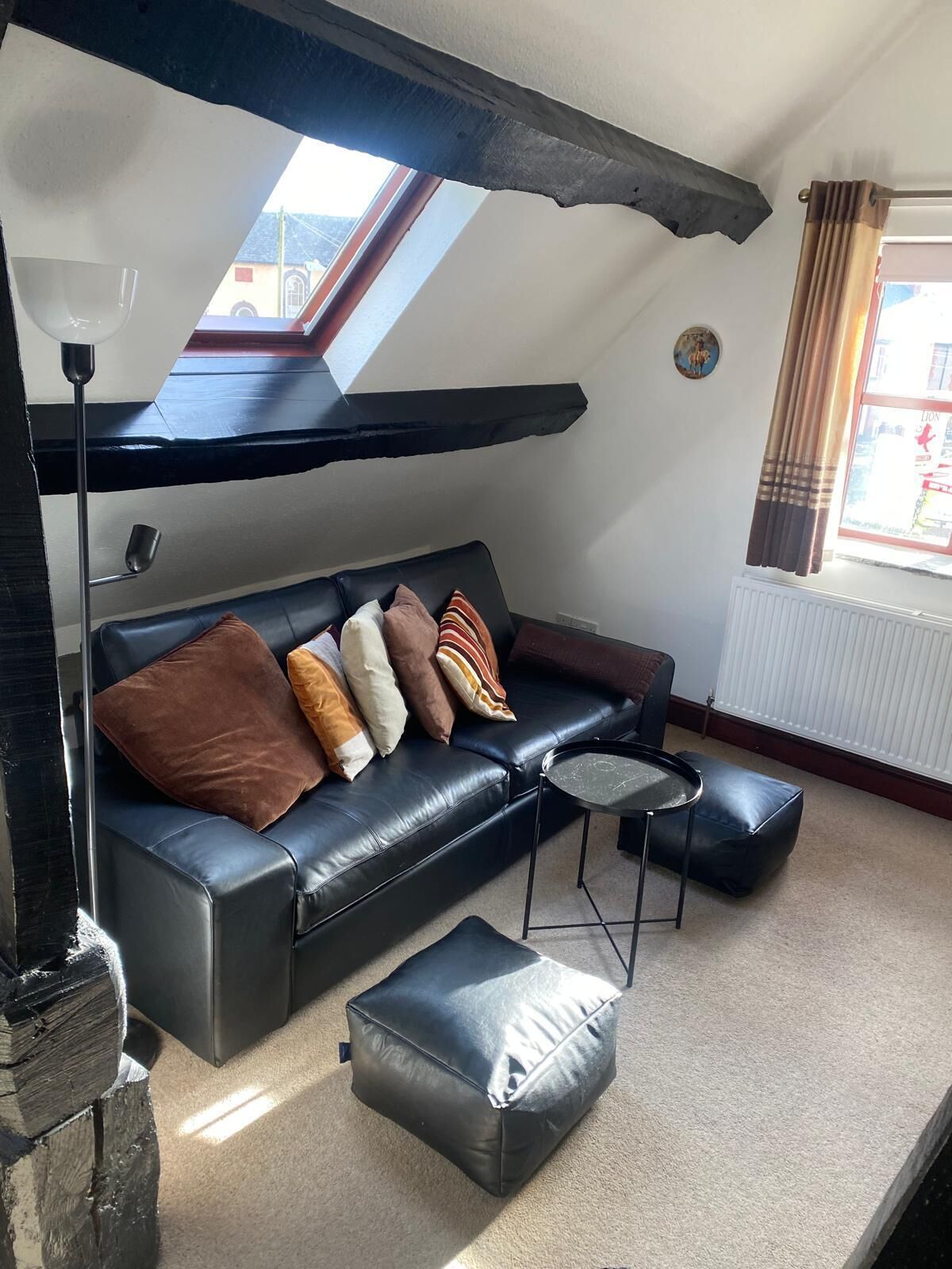 A cozy living room with a black leather sofa, a small round table, and a black pouf. There is a skylight above the sofa and a radiator next to the window.