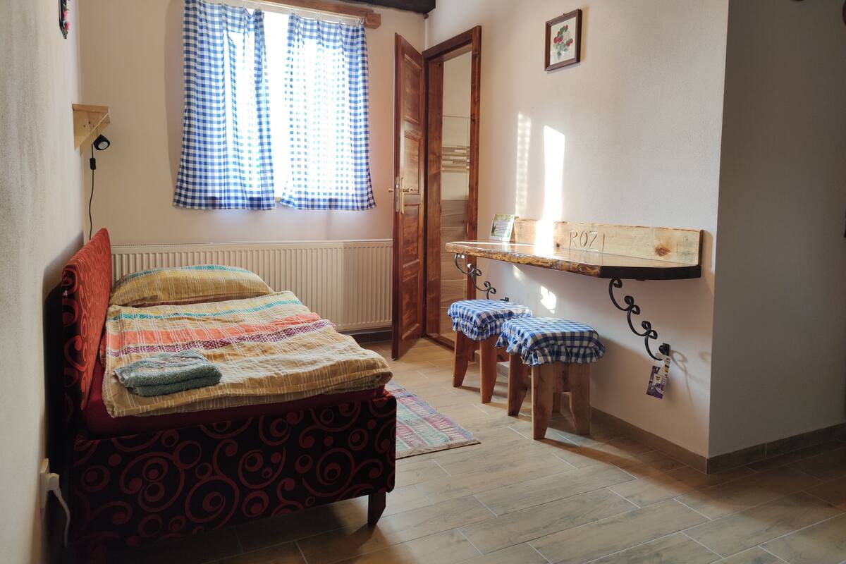Een slaapkamer met een bed, een raam met geruit gordijn, een houten bankje en een klein plankje met naambordje.
