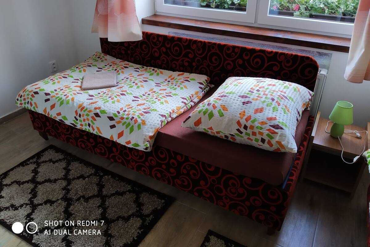 Een bed met beddengoed met een patroon, een raam met uitzicht op groen, en een groene lamp op een nachtkastje.