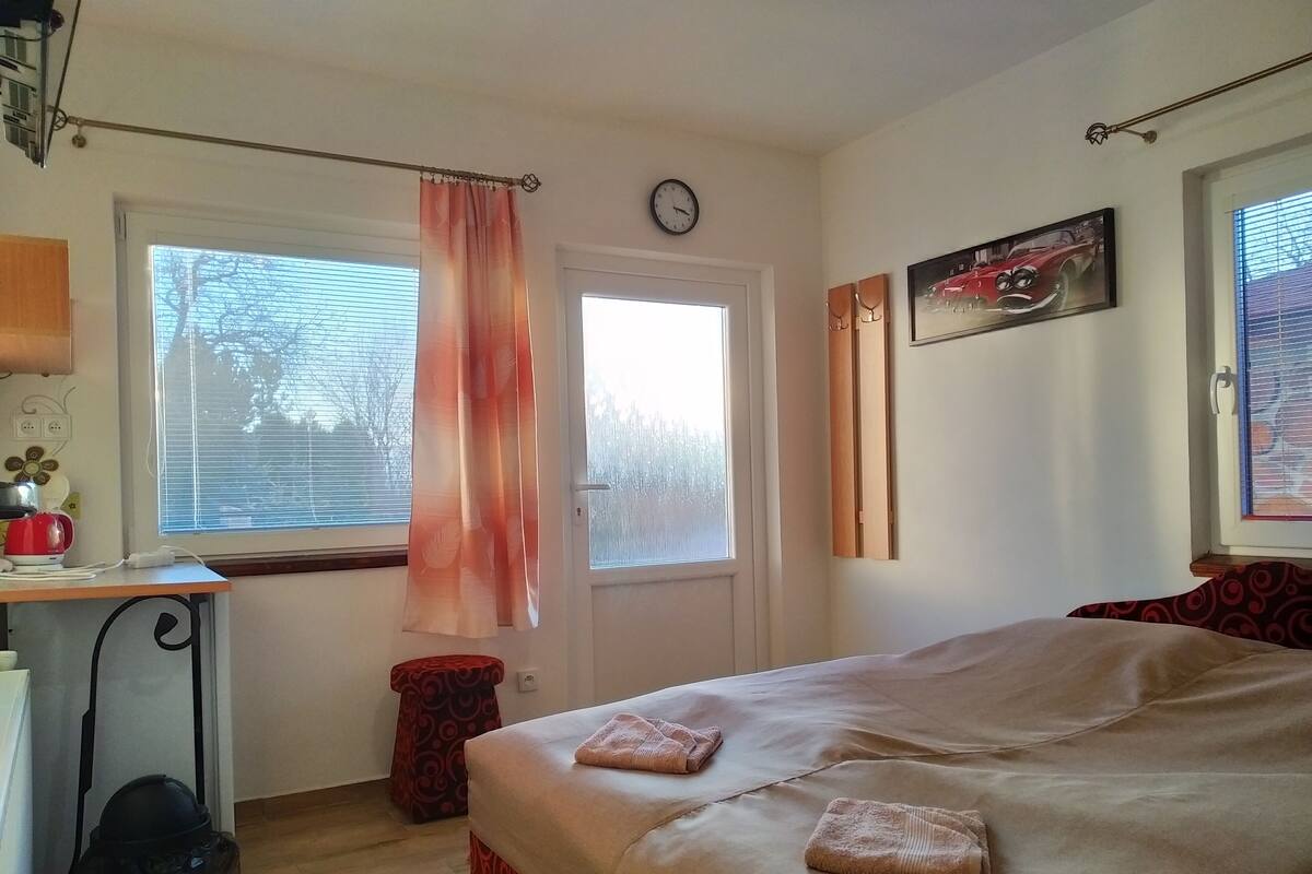 Een slaapkamer met een bed, een bureau, een stoel, een raam met jaloezieën, een deur en een klok aan de muur.