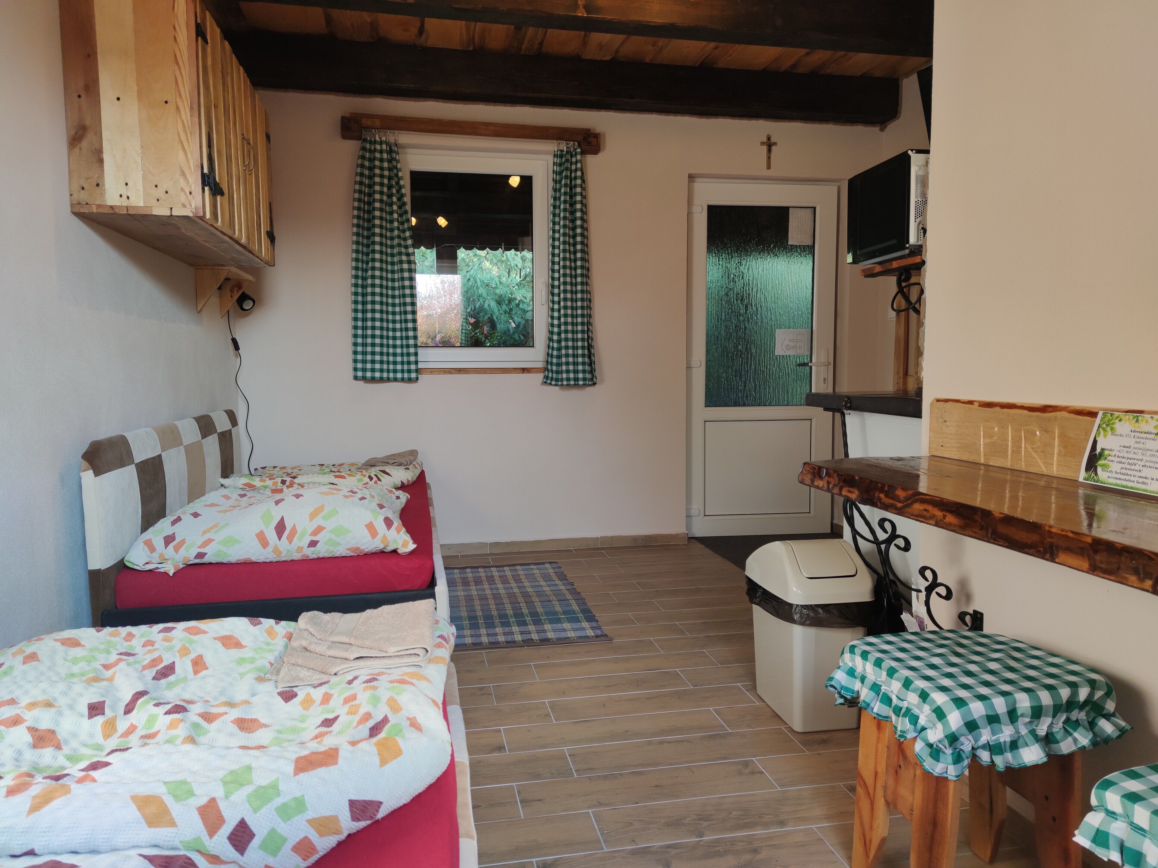 A small, cozy room with two beds, a wooden bench, a small table, and a window with checkered curtains.