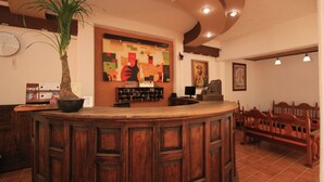 A reception area with a wooden counter, a potted plant, a colorful abstract wall art, and a seating area with wooden chairs.