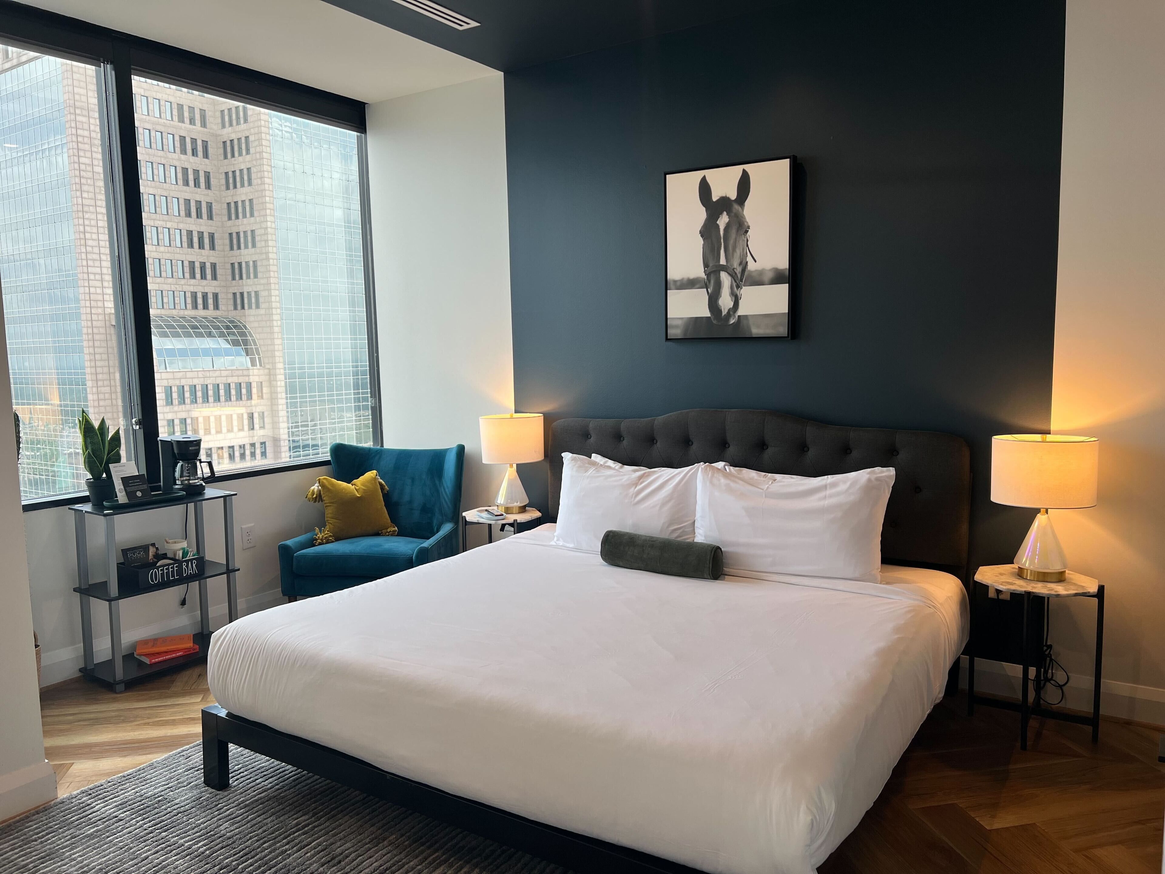 A modern hotel room with a large bed, two bedside lamps, a coffee bar, and a view of the cityscape.