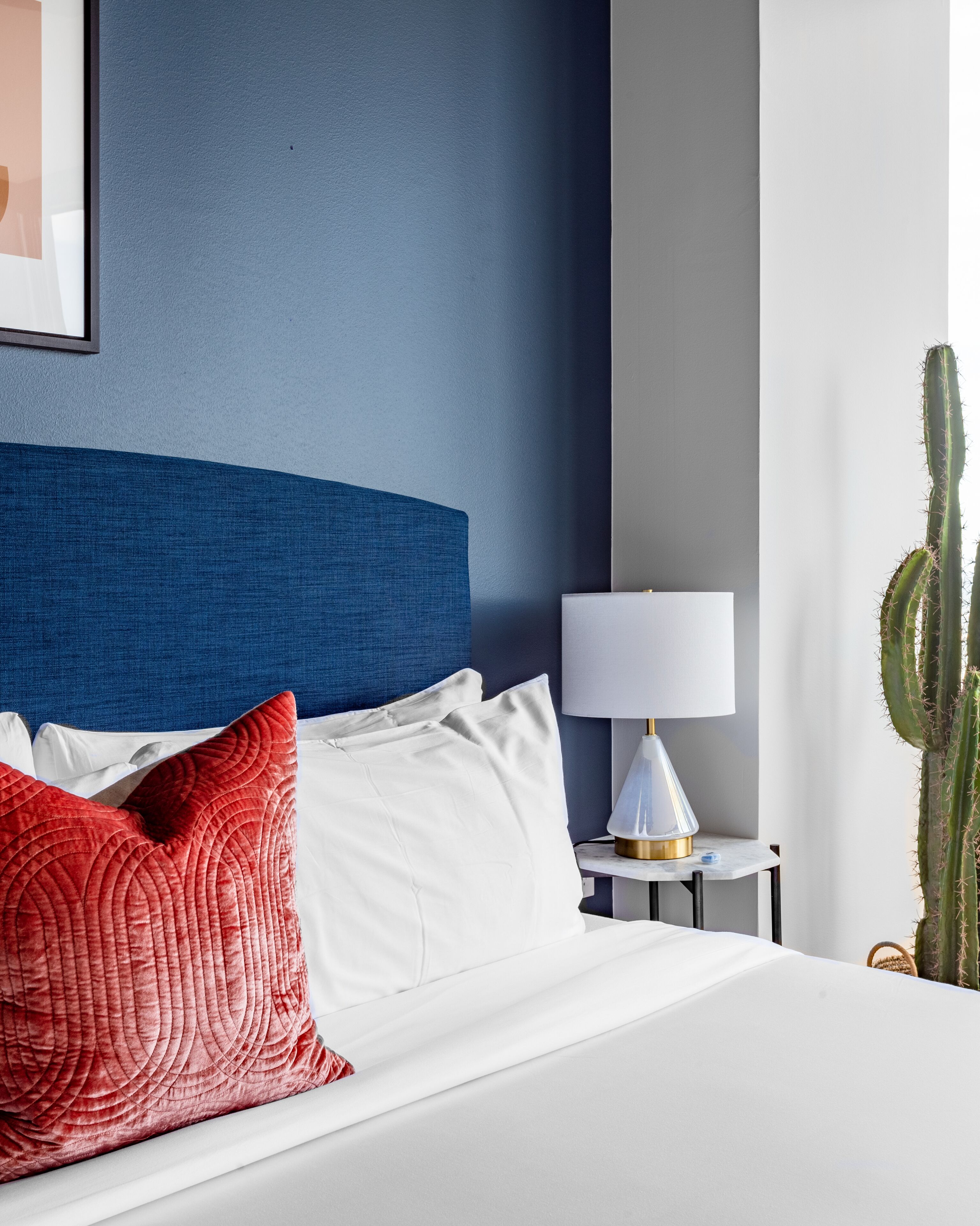 Cama bem arrumada com almofadas brancas e vermelhas, cabeceira azul, mesa de cabeceira branca com abajur e uma planta de cacto.