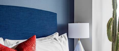 Una cama bien hecha con almohadas blancas y rojas, un cabecero azul, una mesita de noche blanca con una lámpara y una planta de cactus.