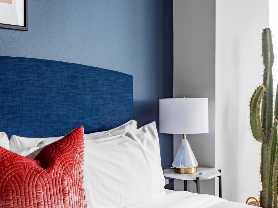 Cama bem arrumada com almofadas brancas e vermelhas, cabeceira azul, mesa de cabeceira branca com abajur e uma planta de cacto.