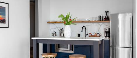 Una cocina moderna con encimera blanca, dos taburetes de madera, un refrigerador de acero inoxidable y un microondas.