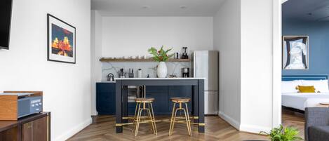 A modern hotel room with a kitchen area, a dining table with stools, a sofa, and a coffee table.