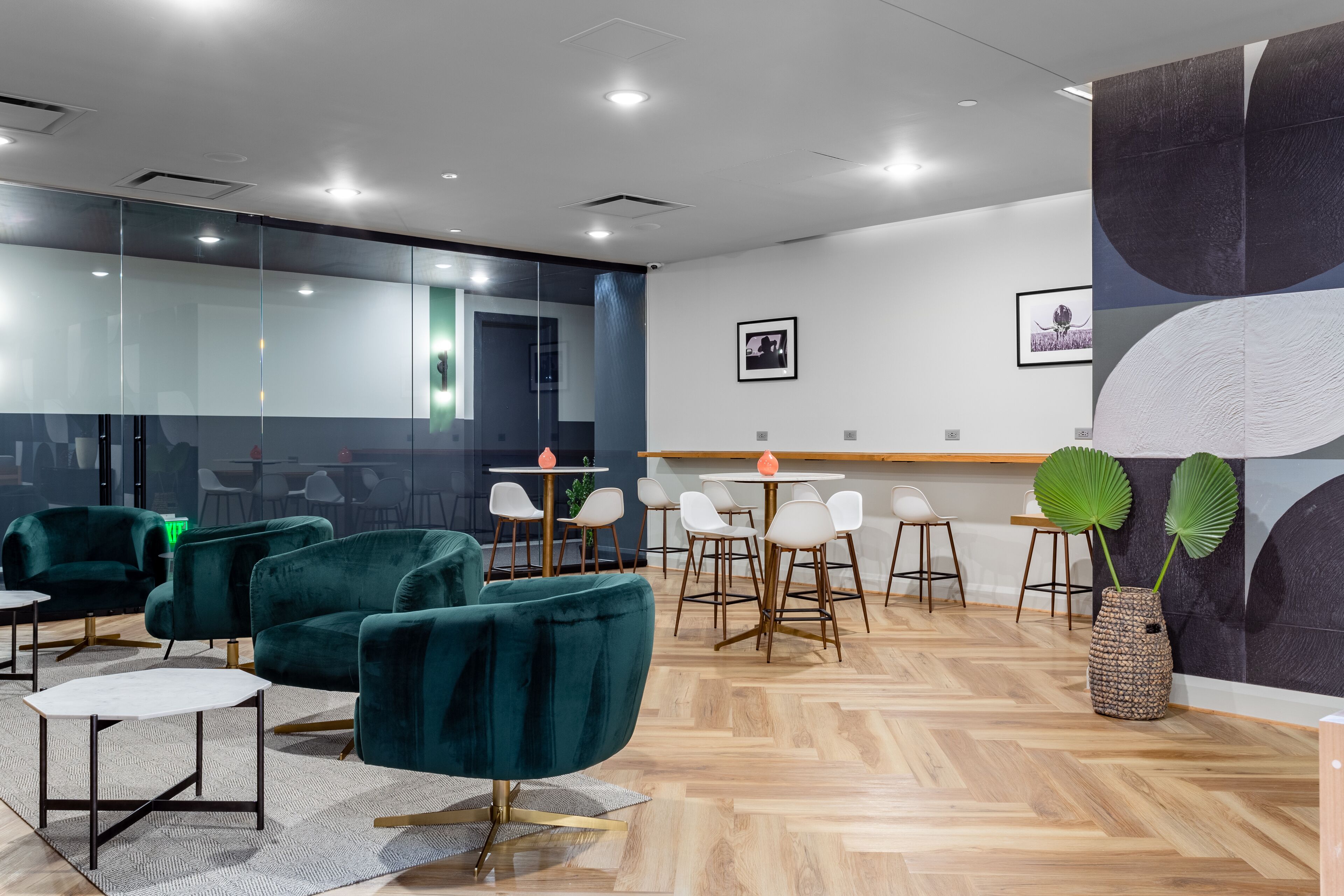 A modern office space with teal upholstered chairs, a wooden herringbone floor, and a glass partition wall.