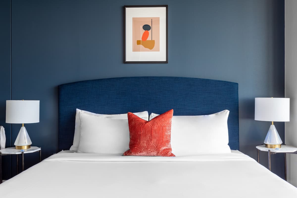 A neatly made bed with white linens and a red accent pillow, flanked by two bedside tables with lamps, set against a dark blue wall with a framed abstract artwork above.
