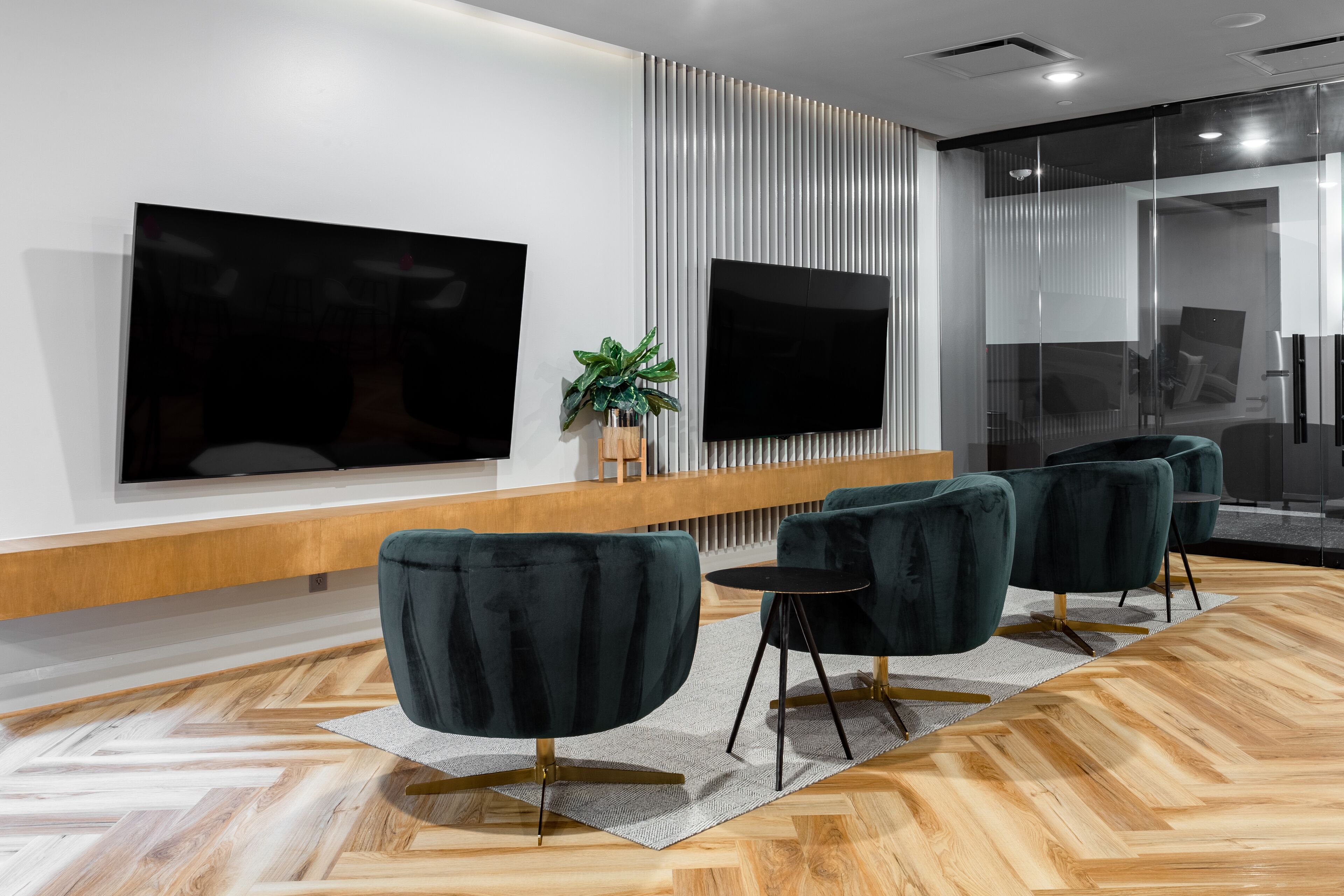 A modern office space with a large flat-screen TV, dark green upholstered chairs, a wooden bench, and a potted plant.
