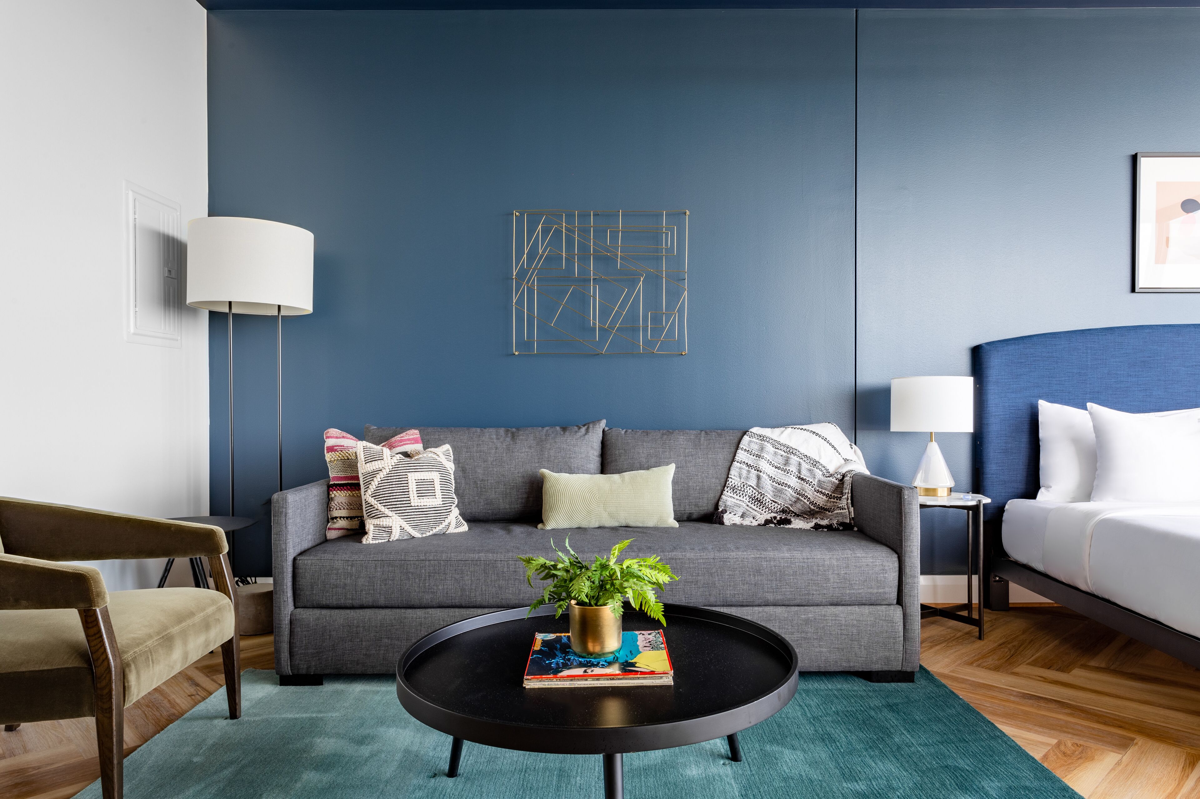 A modern living room with a grey sofa, a round coffee table with a plant, a framed abstract artwork, and a white bed.