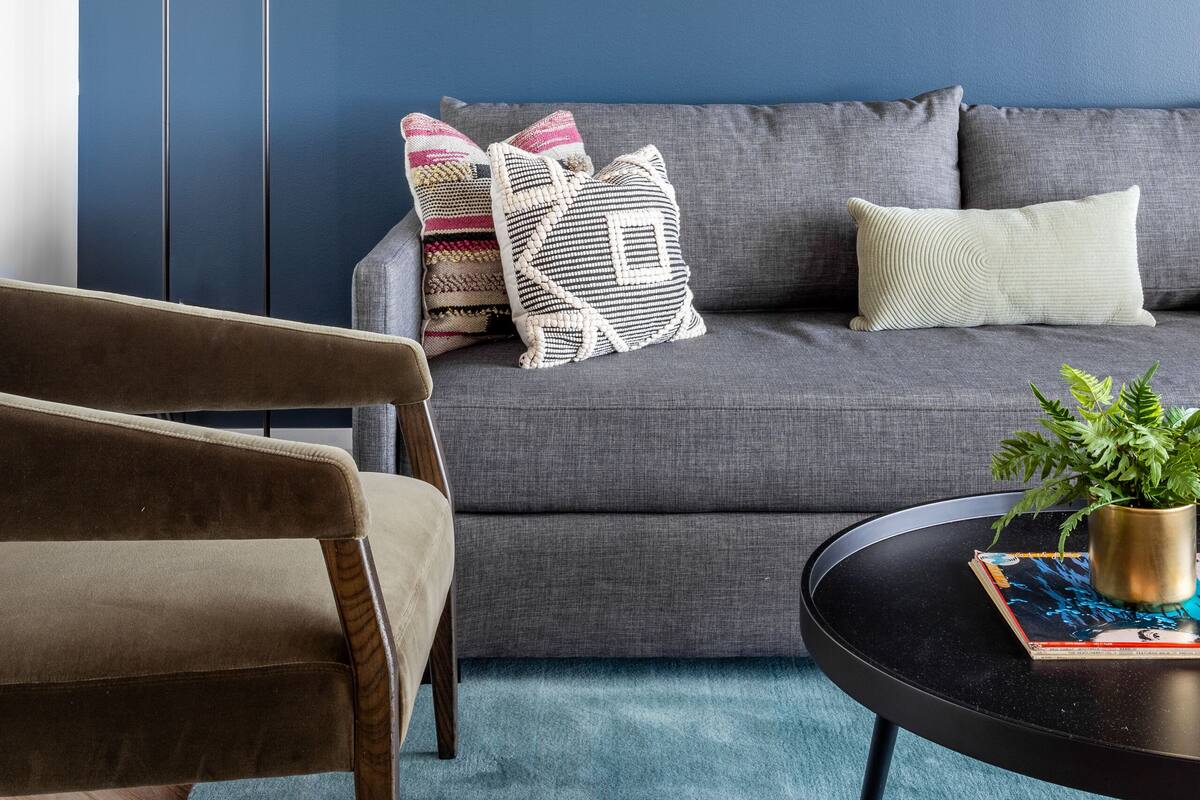 A modern living room with a dark blue wall, a grey sofa, a wooden chair, a floor lamp, and a geometric wall art piece.