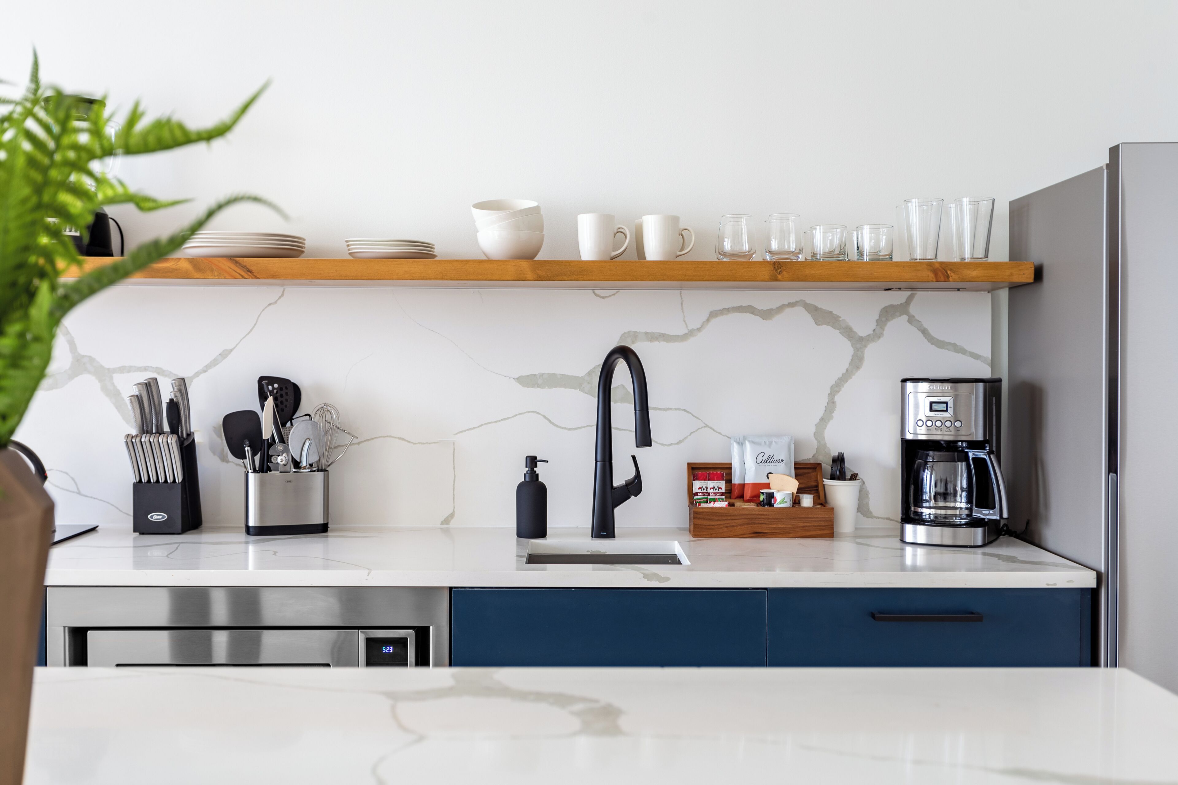 Eine moderne Küche mit weißem Marmorfliesenspiegel, einer Edelstahl-Kaffeemaschine und einem Holzregal über der Spüle.