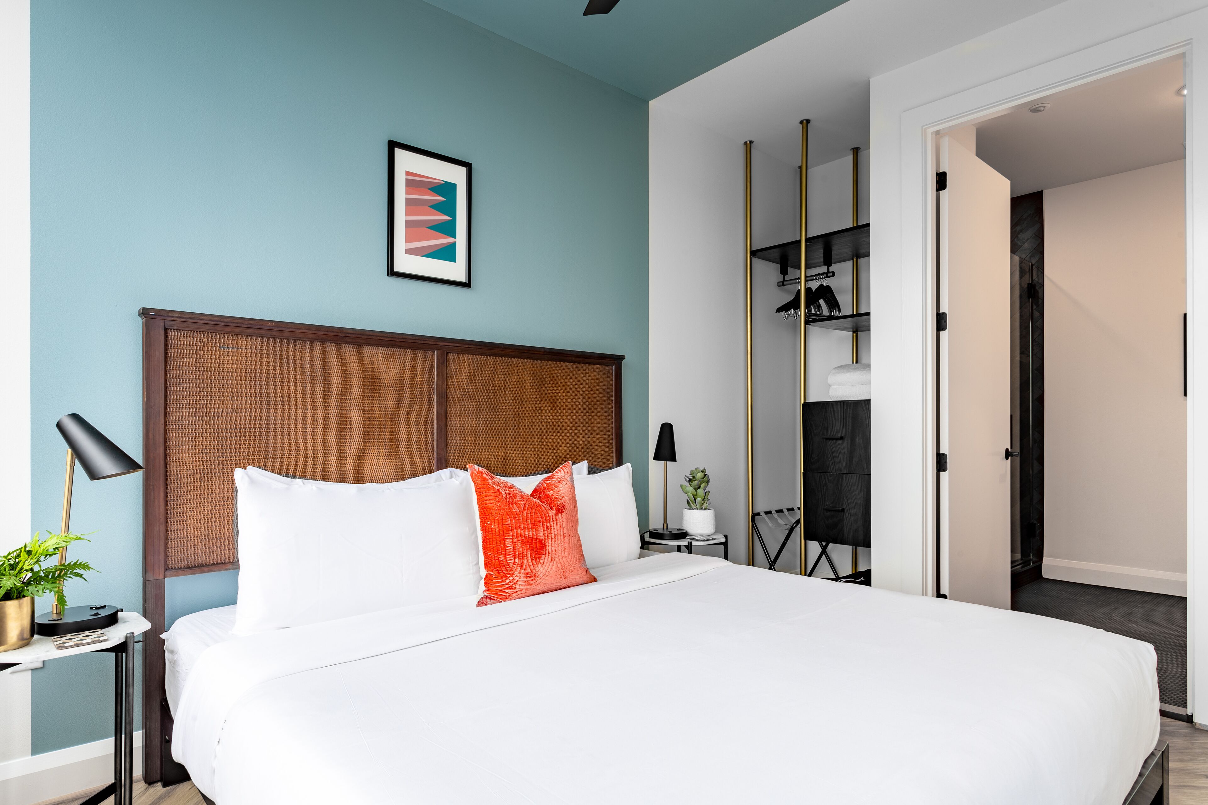 A modern hotel room with a large bed, a wooden headboard, a small bedside table with a lamp, a framed picture on the wall, and a closet with a mirror.
