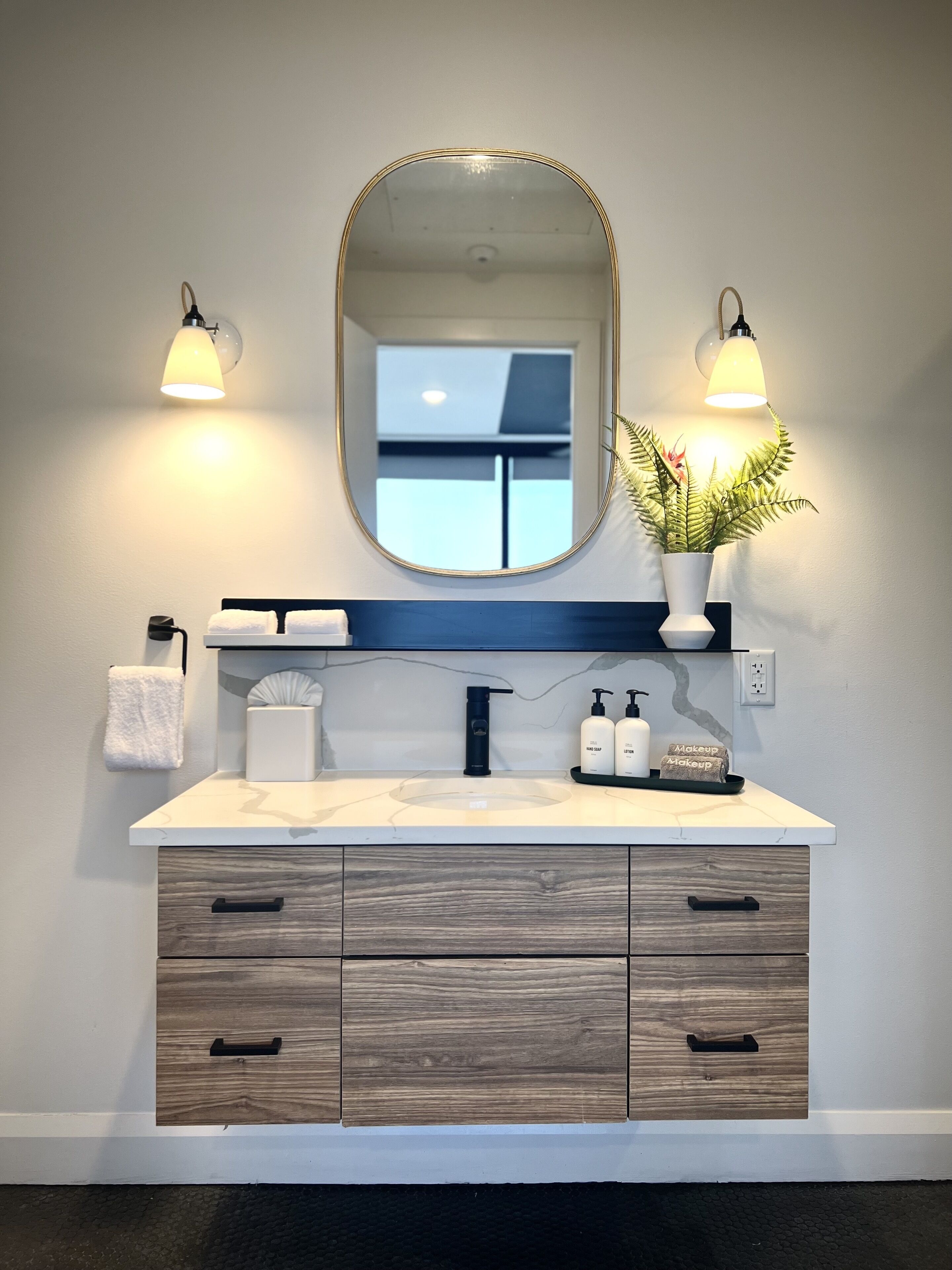 A bathroom with a vanity, two wall-mounted lights, a mirror, a potted plant, a towel, and a soap dispenser.