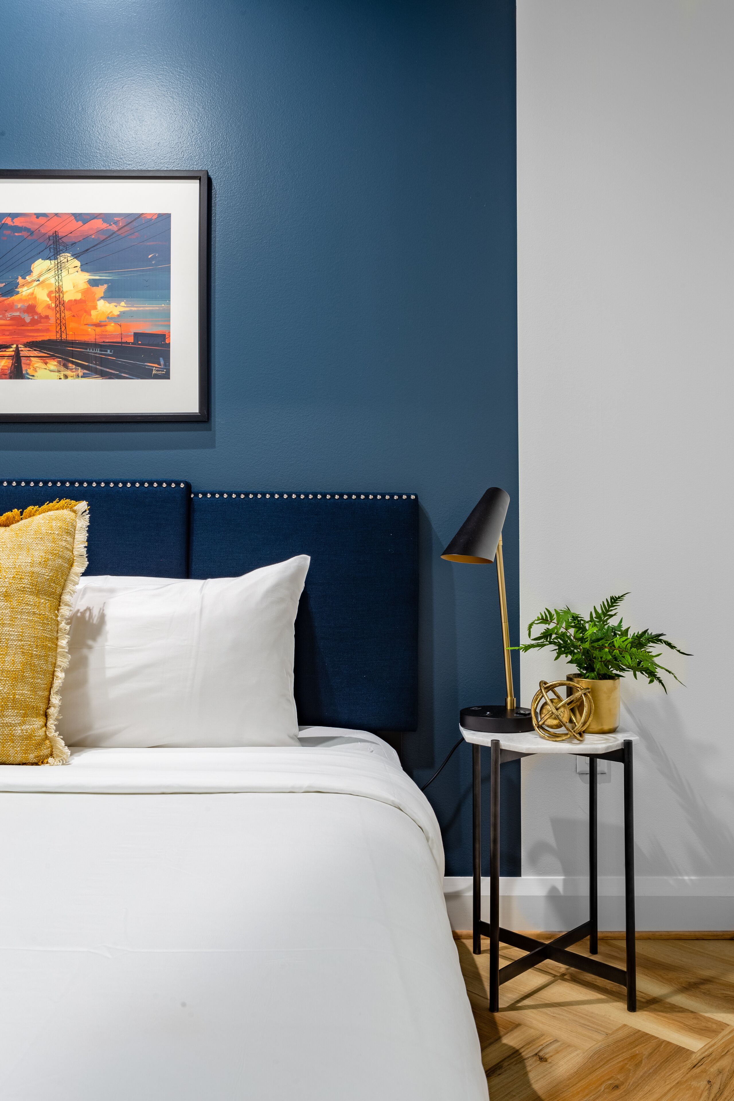Un dormitorio con cama, un cuadro en la pared, una mesita de noche con lámpara y una planta en maceta.