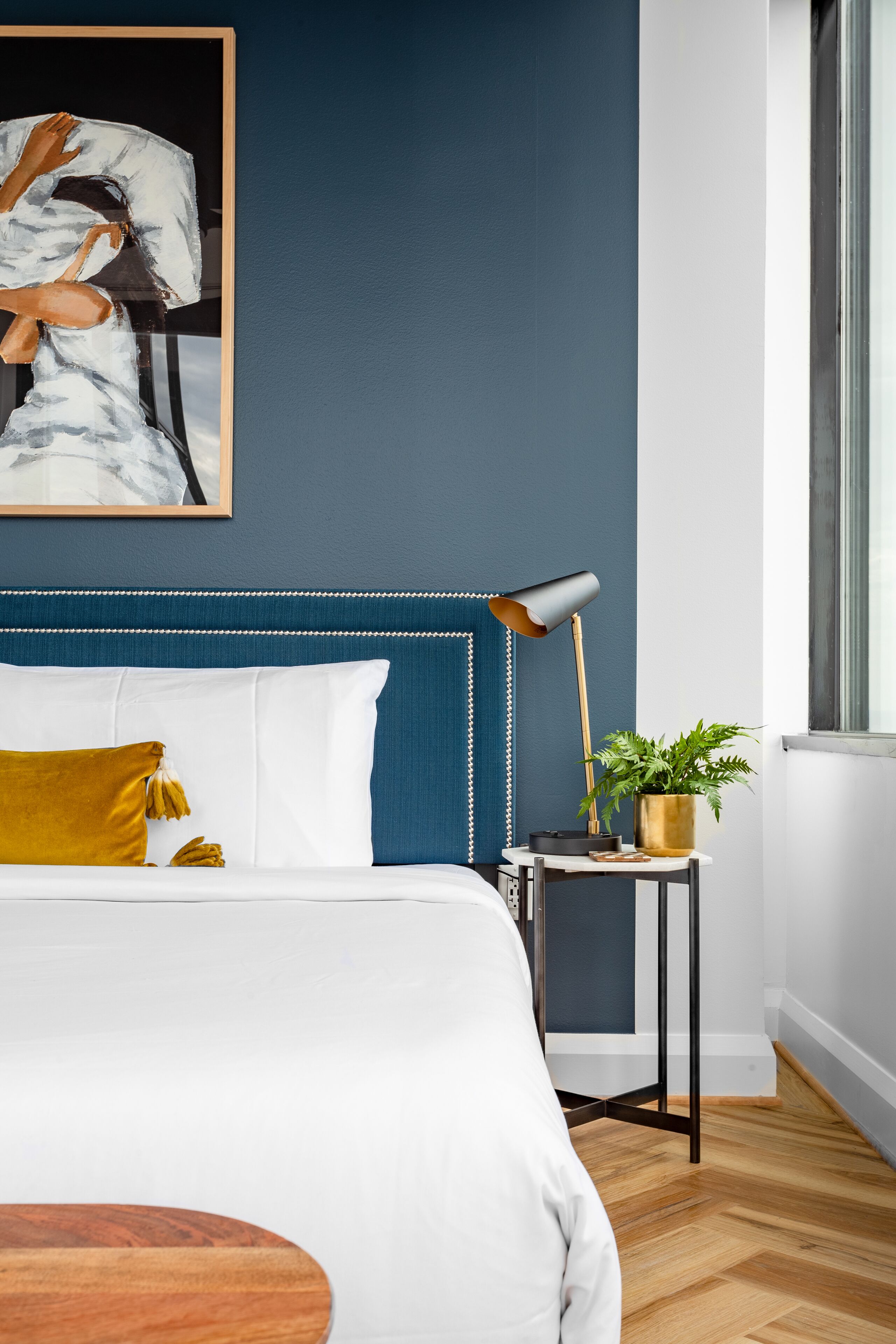 A modern bedroom with a blue accent wall, a bed with white and mustard pillows, a floor lamp, and a small table with a plant.