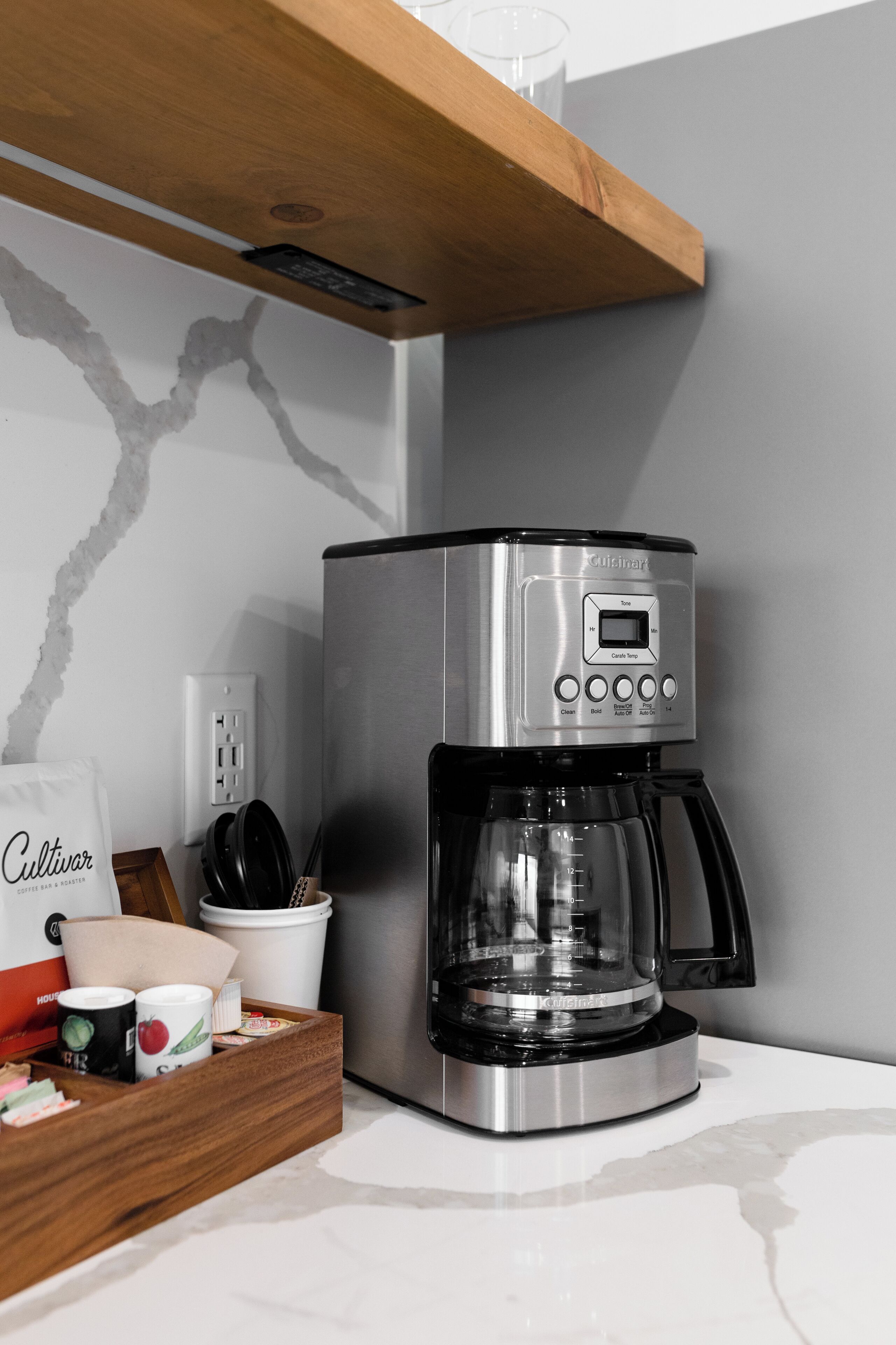 A kitchen with a stainless steel coffee maker, a wooden shelf, a white countertop, and a coffee bag.